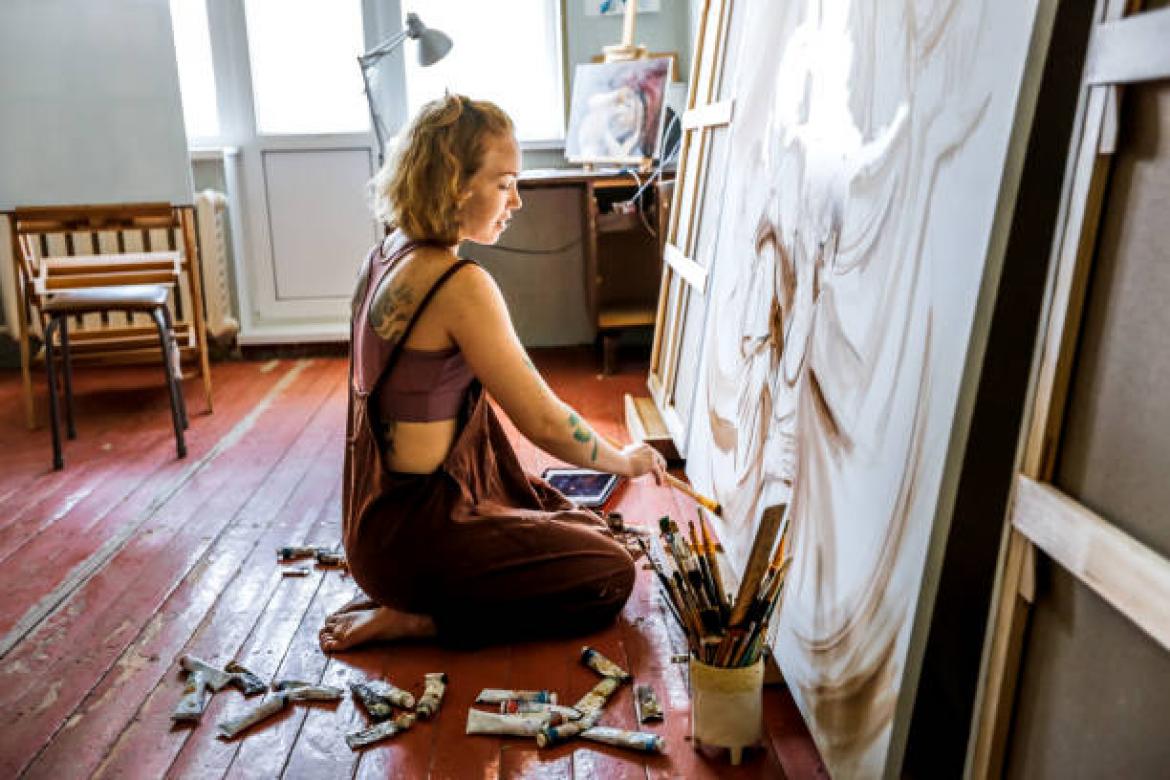 Red-haired female artist with tattoos in linen overalls draws large picture while sitting on floor in her creative workshop