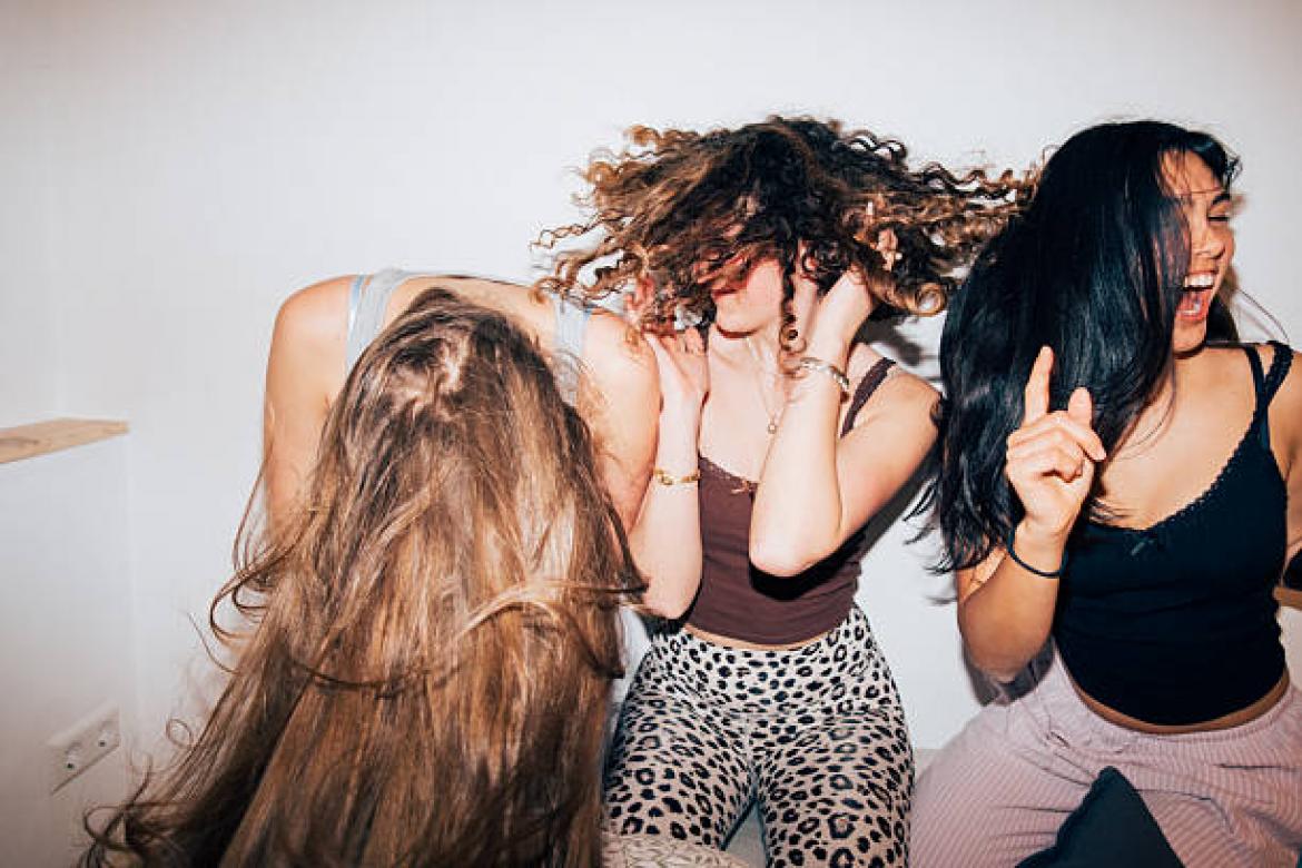 Three women share a lively moment, flipping hair and laughing as they dance in a bright, minimal room. A candid scene of friendship, energy, and carefree youth. Part of a series with video