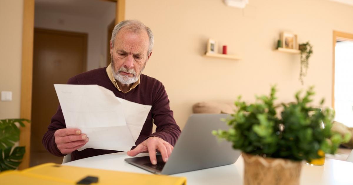 Hombre mayor revisando una factura frente al ordenador en casa.