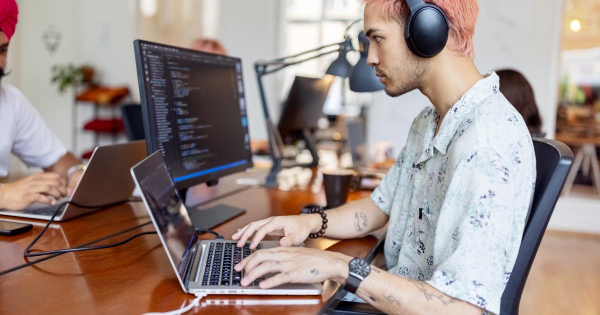 Joven con auriculares trabajando en la computadora en una oficina de startup. Joven profesional de TI trabajando en una oficina de coworking con gente trabajando al fondo.