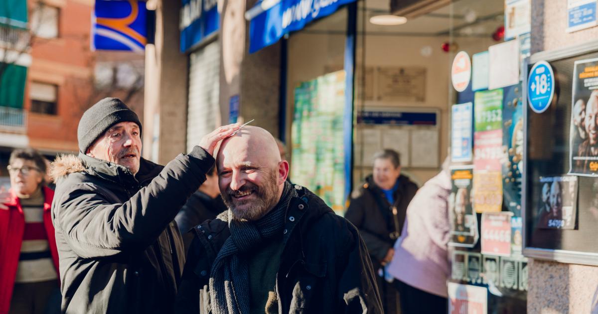 Dos hombres sonríen mientras uno frota un décimo de la Lotería de Navidad en la calva del otro frente a la administración Gaia Loterías en Sant Boi.