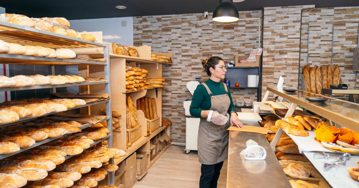 Una trabajadora de panadería entre estantes de panes frescos, manipulando una bolsa de harina en preparación para la siguiente tanda de horneado.