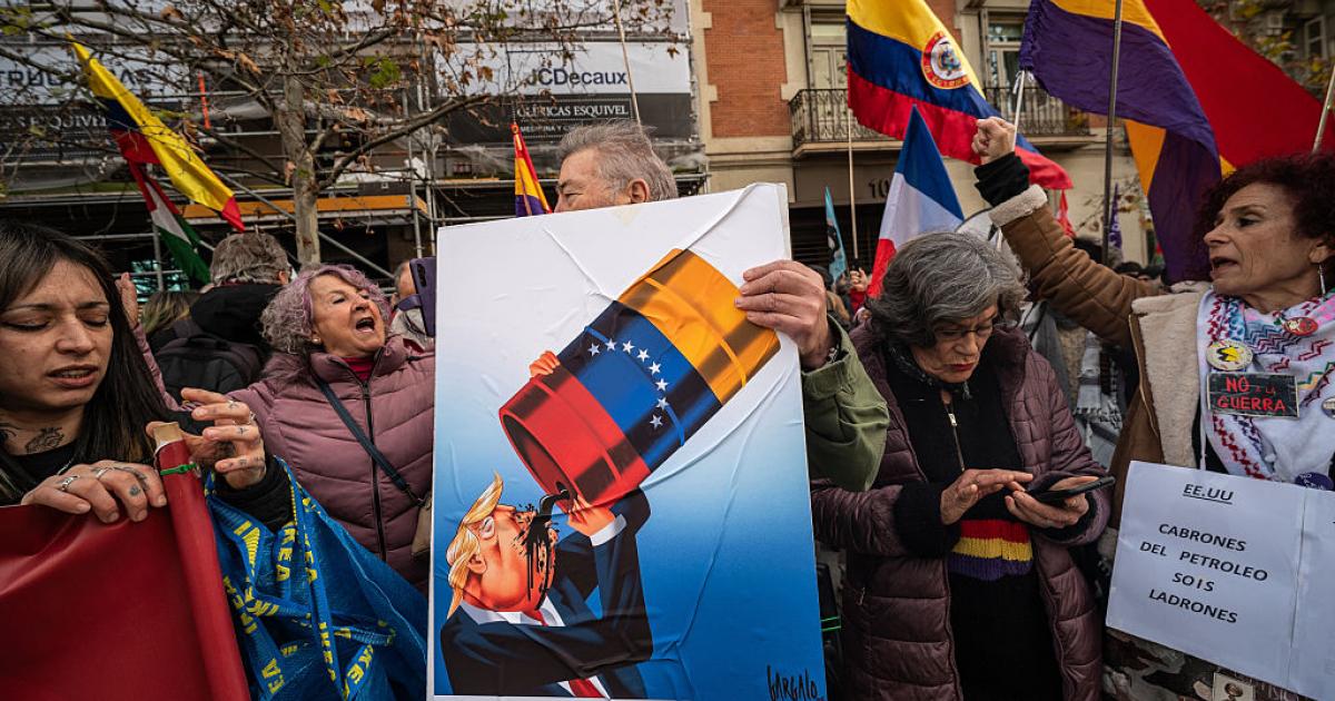 Unos manifestantes ante la embajada de EEUU en Madrid portan un cartel que muestra a Donald Trump bebiendo petróleo venezolano, el 4 de enero de 2026.