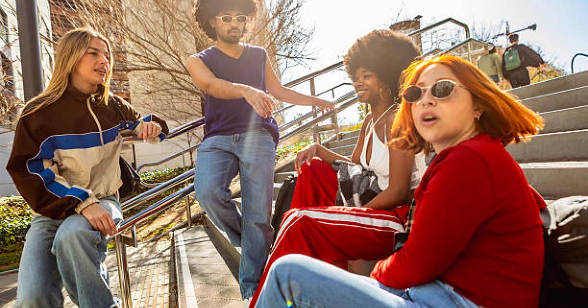 Four friends in casual street style sit on city steps, laughing and chatting in sunny, vibrant urban surroundings, showing diverse gen z friendship and carefree leisure