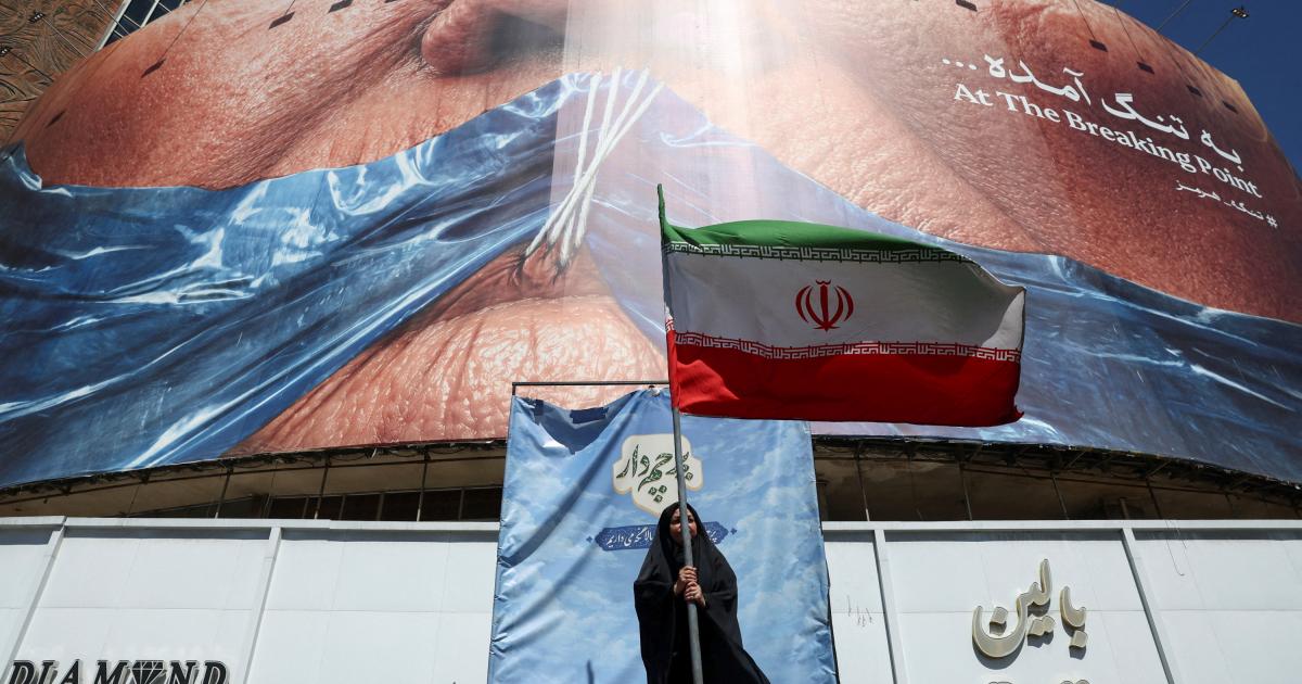 Una mujer sostiene una bandera iraní ant una valla antiestadounidense que muestra al presidente Donald Trump amordazado por el estrecho de Ormuz, en Teherán, el 25 de mayo de 2026.