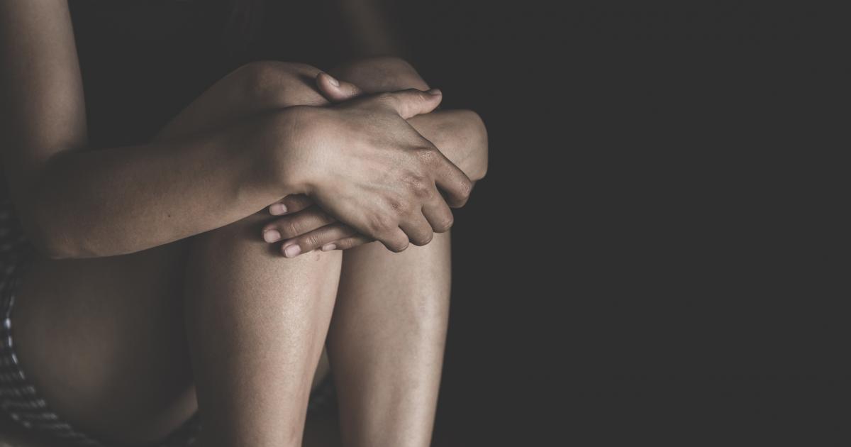 Una mujer abraza sus rodillas con sus brazos, entre las sombras.