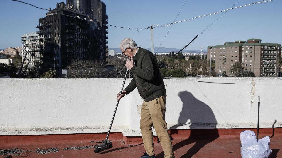 Un vecino de Valencia limpia los restos de ceniza de la azotea, con el edificio incendiado al fondo.