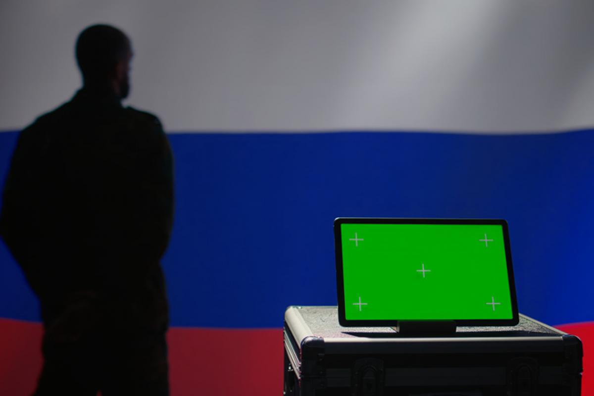 Ilustración de una persona en la sombra con una bandera de la Federación de Rusia al fondo y un portátil.