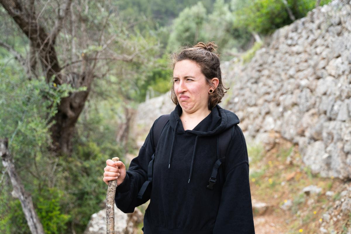 Una joven adulta camina por un sendero forestal con expresión de disgusto. Rodeada de árboles y plantas, sostiene un bastón y una mochila.