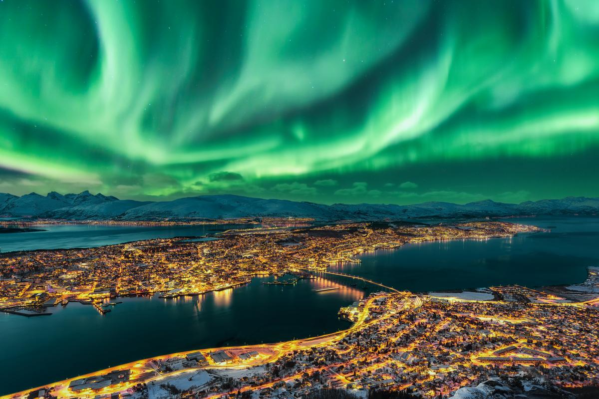 Auroras boreales danzando sobre el horizonte urbano de Tromsø. Panorama de Tromsø y paisaje nocturno invernal con auroras boreales (Aurora Polaris) en una noche fría, con cielo estrellado, luces polares y montañas nevadas en la isla de Kvaloya al fondo.