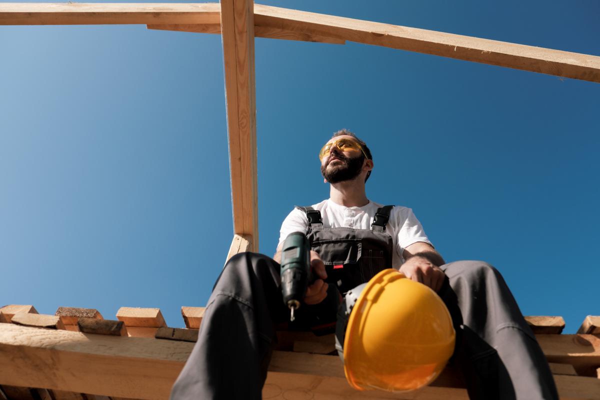 El constructor se sienta en el borde del tejado de la casa de madera, con casco amarillo, overol gris y camiseta blanca. El cielo azul y un día despejado y soleado.