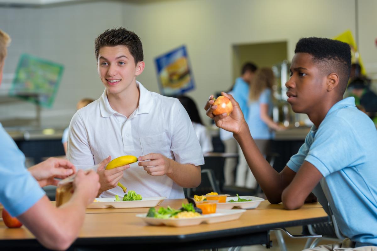 Adolescentes felices y diversos almuerzan juntos en la cafetería de la escuela. Llevan uniforme escolar.