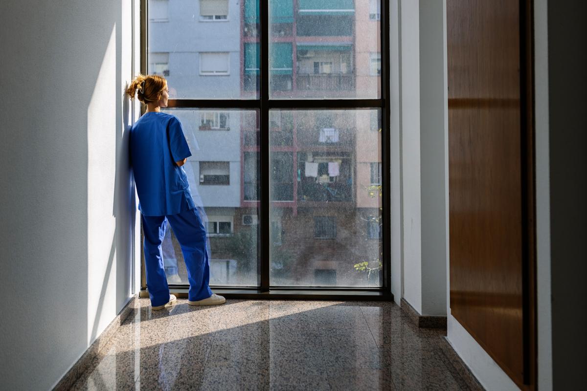 Enfermera de espaldas, con uniforme azul, mirando hacia el exterior desde un pasillo iluminado por la luz natural.