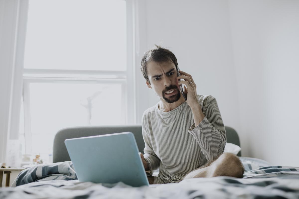 Un hombre enfadado muestra molestia mientras habla por teléfono. Está sentado en su cama con su portátil abierto, y su rostro está animado, mostrando disgusto y frustración.