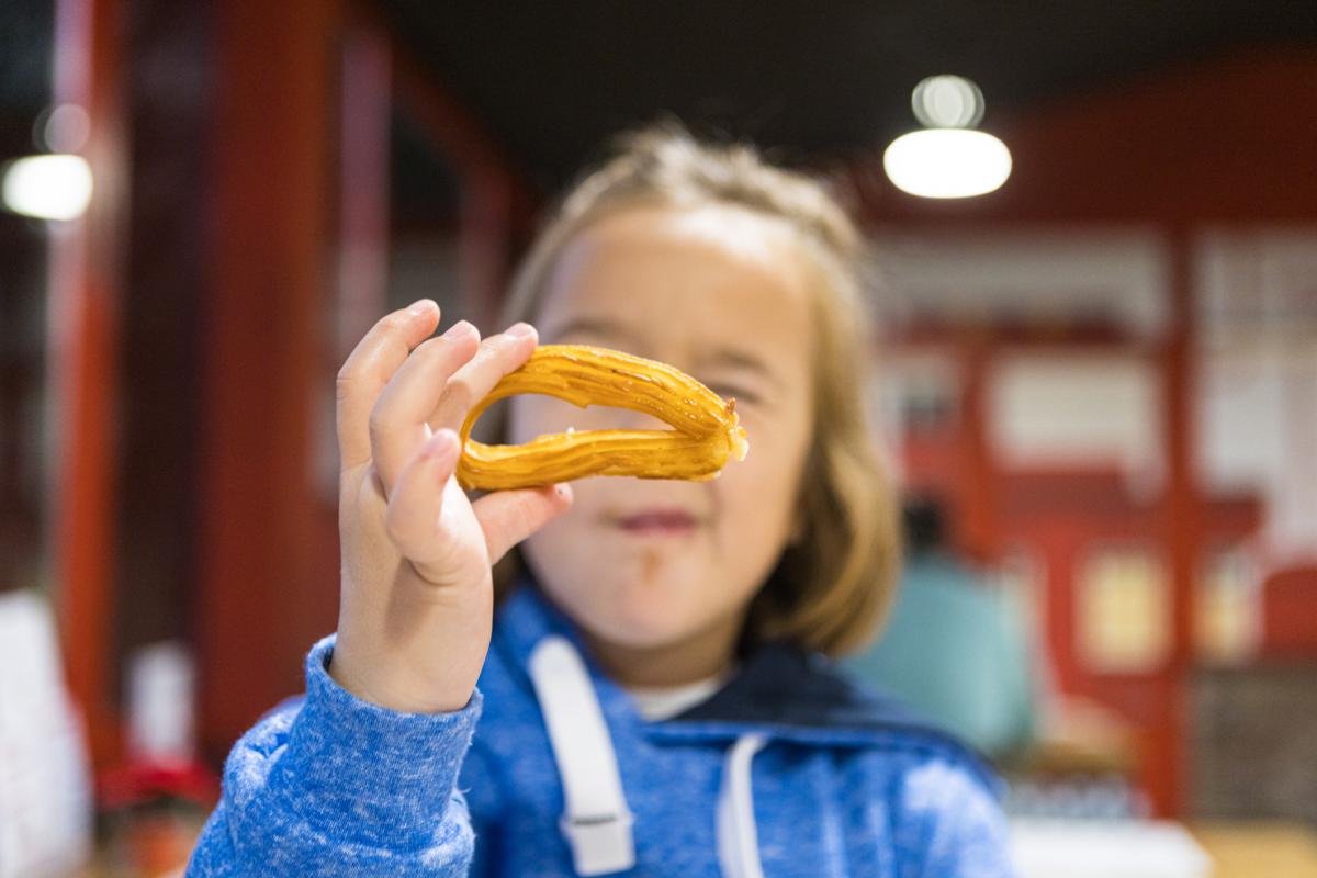 Chica rubia comiendo churros con chocolate sentada en una mesa de cafetería, jugando con uno. Invierno, desayuno, merienda, azúcar y felicidad.