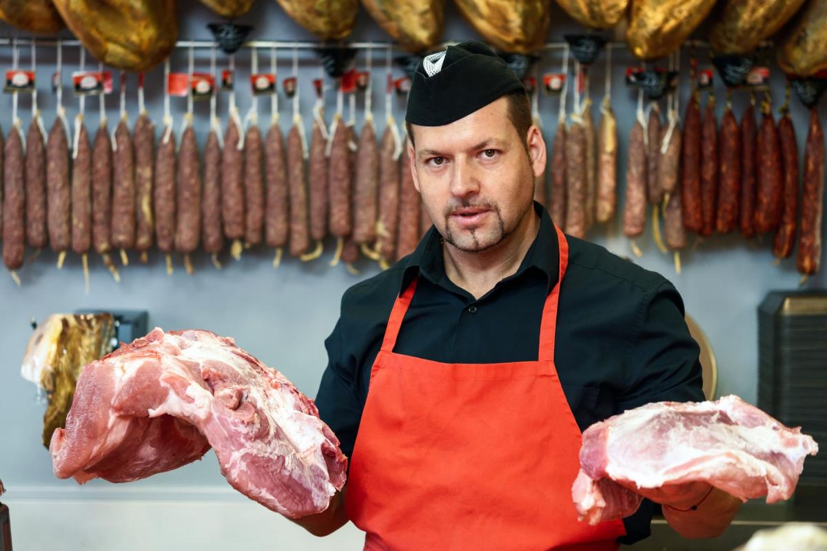 Carnicero de mediana edad sonriendo detrás del mostrador de una carnicería con carne fresca y muchos jamones y embutidos colgando detrás de é