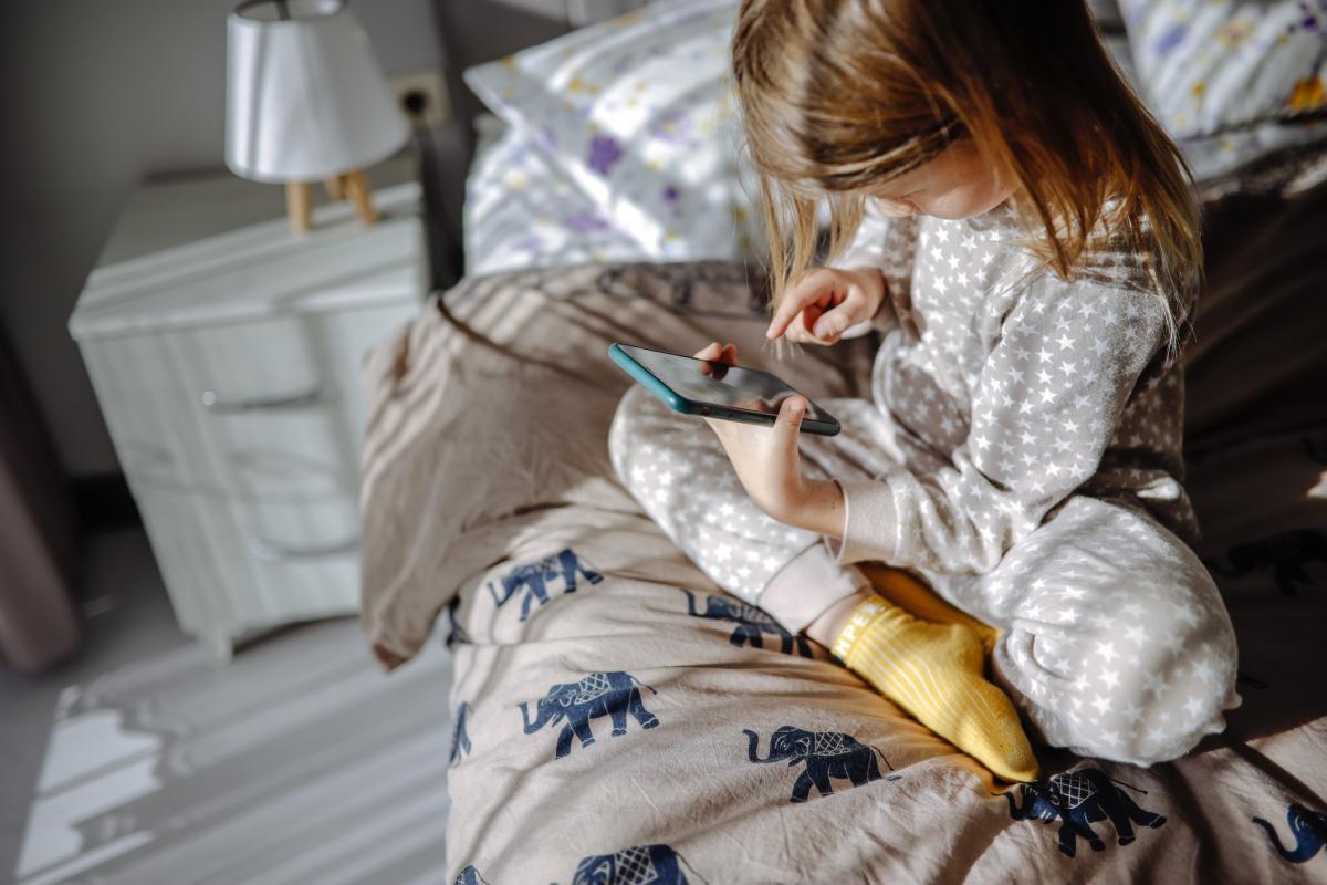 Child in pajamas scrolling on a smartphone while sitting on the bed in the morning, screen time and technology habits.