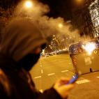 Un encapuchado pendiente del m&oacute;vil mientras un contenedor arde