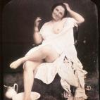 Una mujer sentada en una silla con el camisón colocado de forma que se le puede ver el pecho izquierdo. Tiene el pie derecho en una palangana, como si estuviera lavándoselo, y está sujetando un cigarro ruso. Daguerrotipo estereoscópico color...