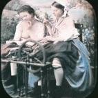 Dos mujeres utilizando una máquina de hilar en un estudio fotográfico con un paisaje campestre de fondo. Una de ellas tiene la blusa desabrochada y se le ve un pecho. Daguerrotipo estereoscópico coloreado a mano. 1850.