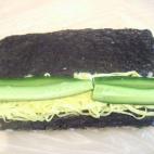 Lo siguiente es darle la vuelta al arroz sobre el alga y colocarlo sobre papel film transparente bien estirado. Sobre el alga vamos colando los hilos de tortilla y los palitos de pepino (paso 1).