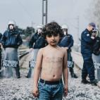 Retrato de un ni&ntilde;o refugiado delante de la polic&iacute;a fronteriza griega con un mensaje de protesta en el pecho &ldquo;no water, no food, open the borders&rdquo; en el campamento de Idomeni, al norte de Grecia. 21/03/2016. Tras meses d...