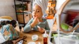 Una niña pequeña sonriente, con una toalla enrollada alrededor de la cabeza, se mira al espejo mientras se aplica crema hidratante en la cara.