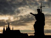 Silueta de la estatua de San Juan Bautista en el Puente de Carlos en Praga, con la Catedral de San Vito al fondo, al atardecer.