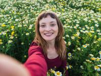 Una mujer sonriendo mientras se toma una fotografía tipo selfie en un campo lleno de margaritas