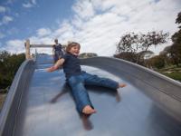 Smiling boy on slide with his mother looking fondly on in background.