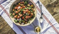 Una colorida y fresca ensaladera con cubos de sandía, tomates cherry, rúcula, rábanos, menta y ralladura de limón. Fotografía tomada bajo el sol radiante en una mesa de picnic en Portugal, durante la primavera. Un pequeño frasco de aderezo, zumo de limón y aceite de oliva acompaña esta ensalada fresca y saludable, ¡lista para comer!