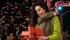 Una mujer abriendo un regalo con cara de sorpresa y un árbol de Navidad de fondo.
