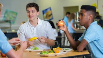 Adolescentes felices y diversos almuerzan juntos en la cafetería de la escuela. Llevan uniforme escolar.