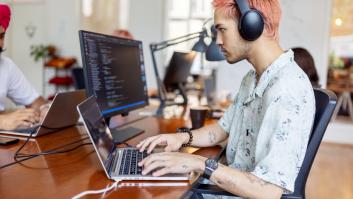Joven con auriculares trabajando en la computadora en una oficina de startup. Joven profesional de TI trabajando en una oficina de coworking con gente trabajando al fondo.