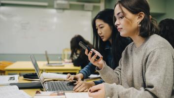 Estudiante de secundaria que usa un teléfono inteligente mientras está sentada junto a un compañero de clase estudiando en una computadora portátil en el aula.