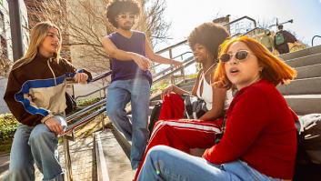 Four friends in casual street style sit on city steps, laughing and chatting in sunny, vibrant urban surroundings, showing diverse gen z friendship and carefree leisure