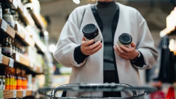 Young Asian woman with shopping cart shopping for cooking sauce in supermarket, reading and comparing the product information of two bottles of cooking sauces. Routine grocery shopping. Making healthier food choices. Consumer awareness concept