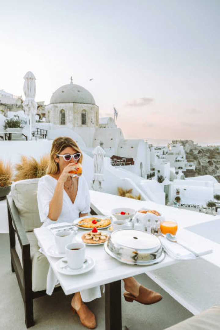 alt="alt="A young tourist woman with a white dress enjoys a healthy breakfast on a luxury resort terrace enjoying egg pancakes, and drinking orange juice, tea, and coffee while taking in the stunning Aegean sea view of Oia or Ia Town, Santorini, Greek islands, Cyclades, and Greece.""