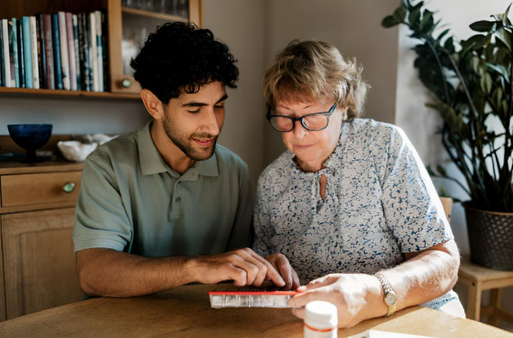 alt="alt="Un enfermero joven, de rodillas, ayudando a una mujer mayor a organizar sus pastillas""
