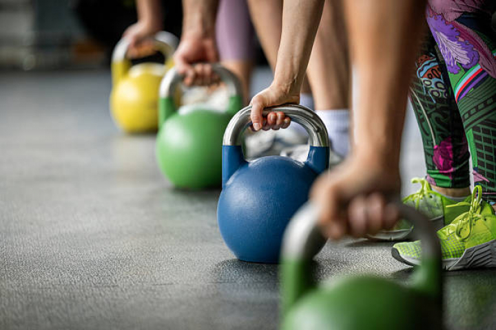 alt="alt="Multiple hands grip kettlebells of different colors, lined up on the floor. The detail shot emphasizes strength, coordination, and functional fitness equipment.""