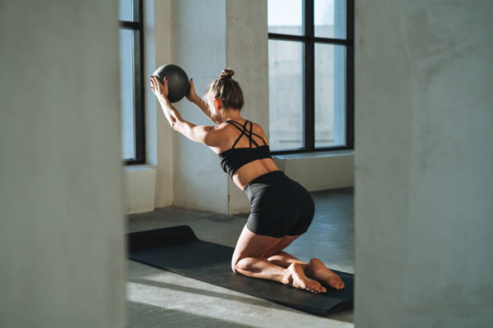 alt="alt="Young slim woman in sportswear doing Pilates stretching on mat with ball in modern studio""