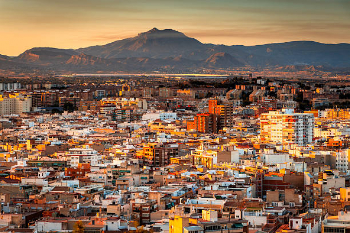 alt="alt="Elevated vantage point depicting the cityscape of the seaside city of Alicante in southern Spain at sunset. The city is bathed in warm light from the dazzling sunset.""