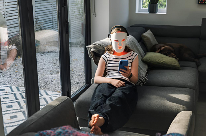 Una adolescente usa su mascarilla facial LED roja (terapia de luz roja) mientras se relaja en el sofá de su casa y mira su móvil