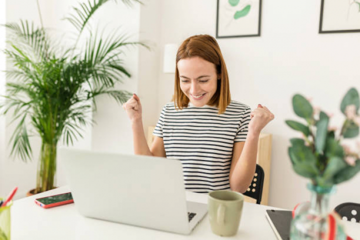 alt="alt="Cheerful young woman celebrating her achievement while reading good news on laptop from home office - Smiling female expressing excitement working at home""