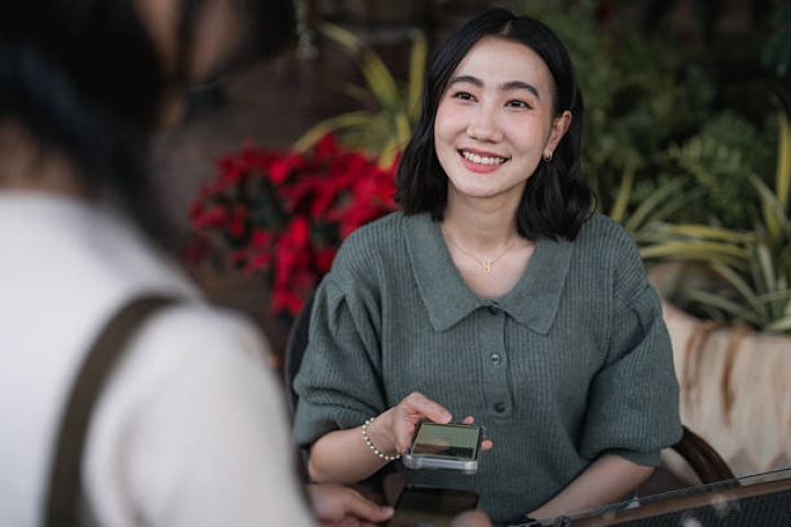 alt="alt="Confident young woman taping her phone against the waitress's device for a quick mobile payment, showcasing the rise of cashless convenience, contactless tech, and seamless digital transactions in today's connected lifestyle""
