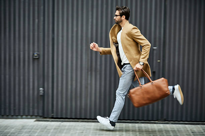 alt="alt="Full length of businessman running on sidewalk. Male professional is carrying bag by building. He is wearing smart casuals in city.""