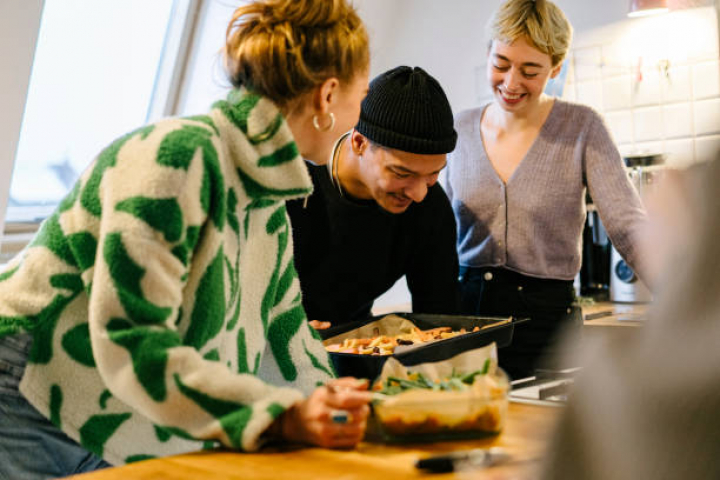 alt="alt="Landscape format of three friends standing together in the kitchen looking happy, he puts a baking tray of vegetables in the oven""