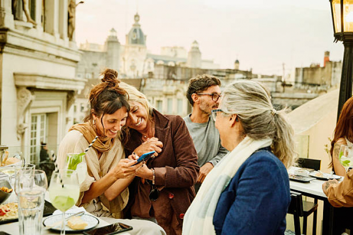 alt="alt="Medium shot of senior mother and adult daughter looking at smart phone and laughing during outdoor party on city rooftop restaurant""