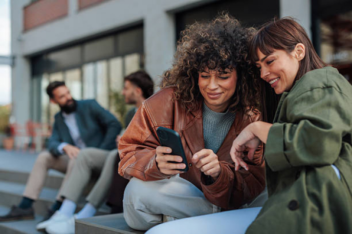 alt="alt="Two young businesswomen are sitting on steps outside their office building, looking at a smartphone and smiling""