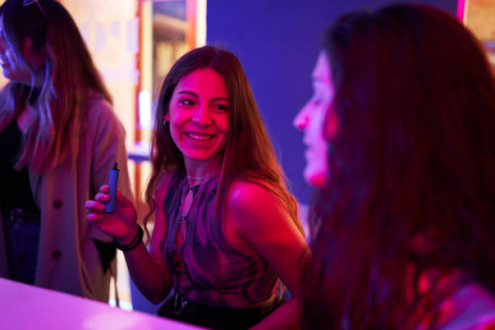 alt="alt="Woman sitting at the bar of a discotheque with an electronic cigarette next to friends""